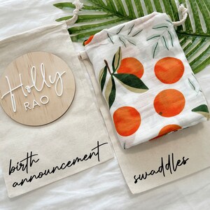 May include: Two white canvas bags with black text. One bag has a wooden circle with the text "Holly Rao" and the text "birth announcement". The other bag has a white muslin swaddle with an orange citrus print and the text "swaddles".
