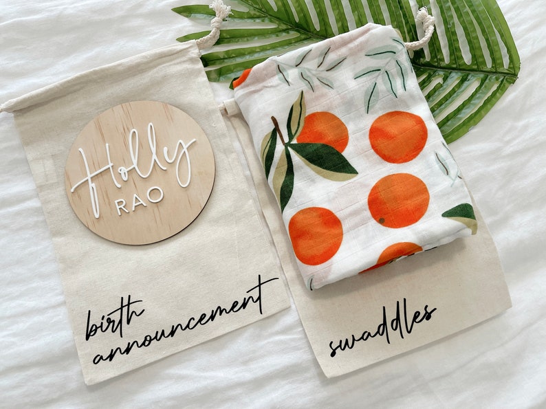 May include: Two white canvas bags with black text. One bag has a wooden circle with the text "Holly Rao" and the text "birth announcement". The other bag has a white muslin swaddle with an orange citrus print and the text "swaddles".