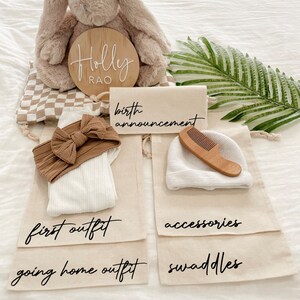 May include: A set of four beige canvas bags with black text labels. The bags are labeled "birth announcement", "first outfit", "going home outfit", and "swaddles". The bags contain a stuffed bunny, a headband, a white onesie, a white hat, a wooden comb, and a few swaddles. The bags are arranged on a white surface with a wooden sign that reads "Holly Rao".
