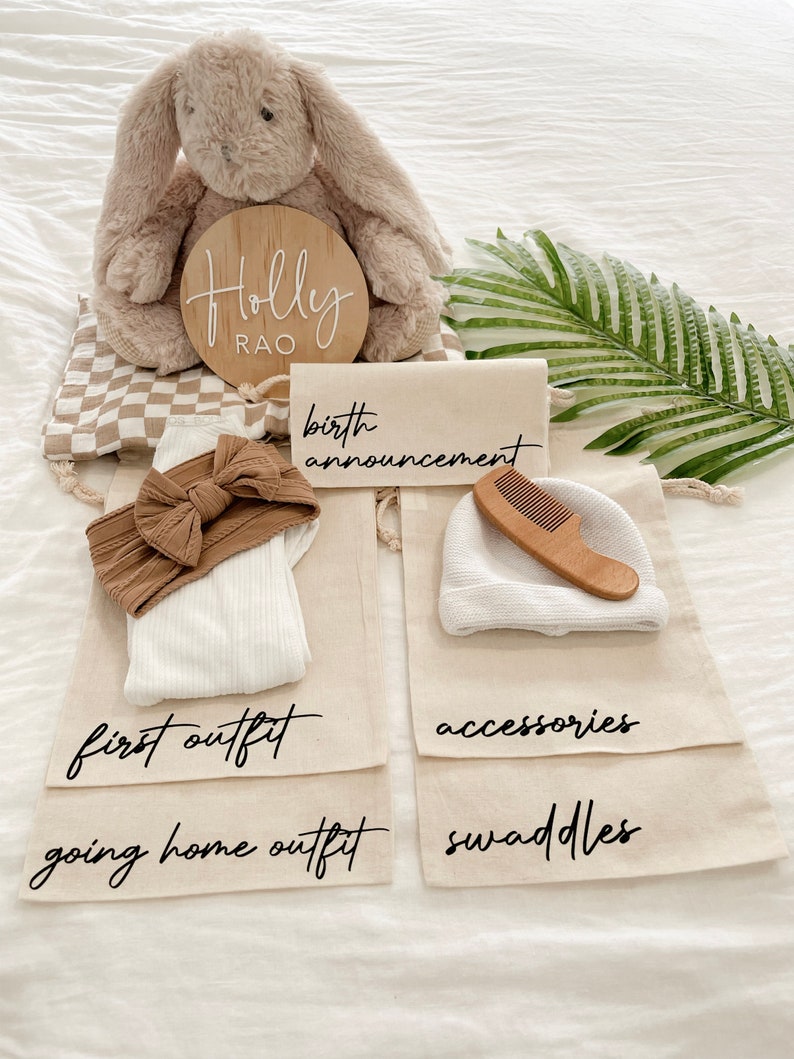May include: A set of four beige canvas bags with black text labels. The bags are labeled "birth announcement", "first outfit", "going home outfit", and "swaddles". The bags contain a stuffed bunny, a headband, a white onesie, a white hat, a wooden comb, and a few swaddles. The bags are arranged on a white surface with a wooden sign that reads "Holly Rao".