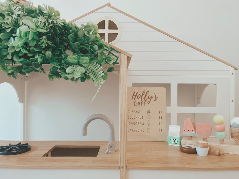 May include: A wooden play kitchen with a sign that reads "Holly's Cafe" and a menu listing prices for cupcakes, doughnuts, ice cream, and babyccinos. The kitchen has a sink, a stovetop, and a window. There are toy ice cream cones and a milk carton on the counter.