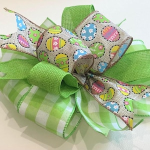 May include: A decorative Easter bow featuring multiple layers of ribbon in green and white gingham, solid green, and a burlap-like ribbon printed with colorful Easter eggs. The bow is a festive decoration.