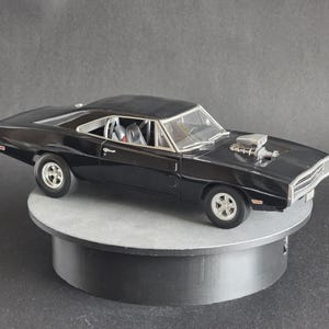 May include: A black die-cast model car with a silver engine component on the hood. The car is on a rotating platform. The model car is approximately 10 inches long.