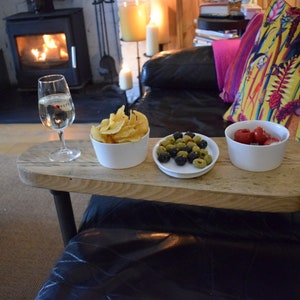 May include: A wooden serving tray with a black metal stand sits on a black leather sofa. The tray holds three white bowls filled with snacks: potato chips, olives, and cherry tomatoes.