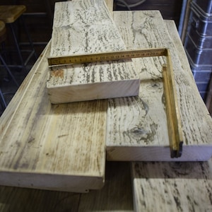 May include: A stack of reclaimed wood planks with a weathered white finish. The planks are arranged in a staggered pattern and have a rustic appearance. A wooden ruler is placed on top of the planks, with the numbers 1-22 visible.