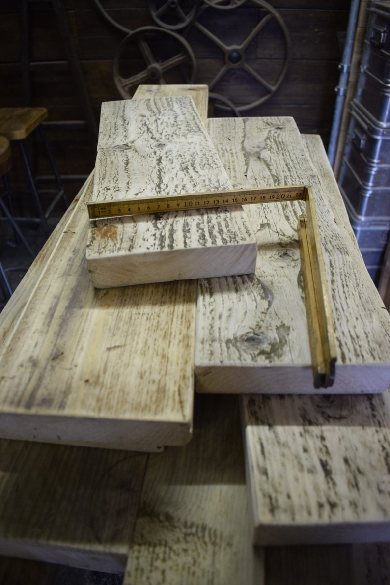 May include: A stack of reclaimed wood planks with a weathered white finish. The planks are arranged in a staggered pattern and have a rustic appearance. A wooden ruler is placed on top of the planks, with the numbers 1-22 visible.