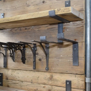 May include: A wall-mounted display featuring a horizontal wooden shelf supported by dark metal brackets. Below the shelf, four distinct metal shelf brackets are attached to the wooden panel, showcasing various ornate and industrial designs.