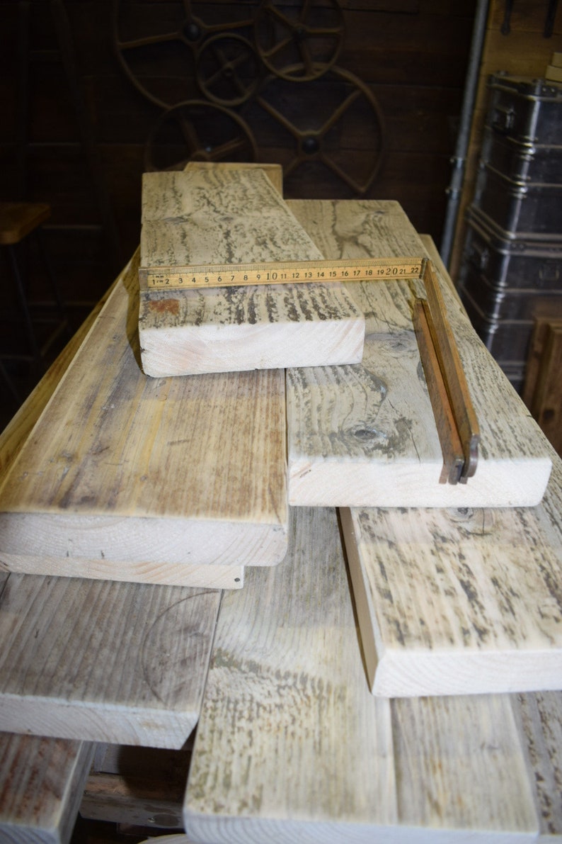 May include: A stack of reclaimed wood planks, some with a weathered gray finish, others with a natural wood color. A wooden ruler is placed on top of the planks, with the numbers 1 through 22 visible.