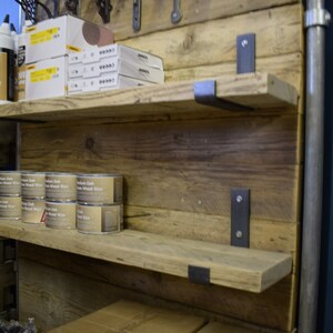 May include: Rustic wooden shelves with metal brackets, mounted on a reclaimed wood wall. The shelves are filled with various items, including cans of wood wax and boxes of supplies.