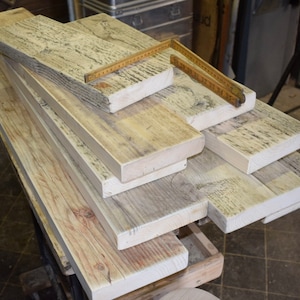 May include: A stack of reclaimed wood planks, some with a weathered gray finish, others with a natural wood color. The planks are arranged in a staggered pattern, with the top plank partially overlapping the one below it. A wooden ruler is lying on top of the planks.
