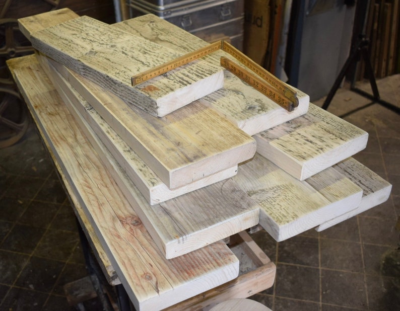 May include: A stack of reclaimed wood planks, some with a weathered gray finish, others with a natural wood color. The planks are arranged in a staggered pattern, with the top plank partially overlapping the one below it. A wooden ruler is lying on top of the planks.