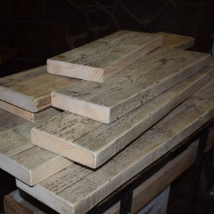 May include: A stack of reclaimed wood planks, showing the natural grain and texture of the wood. The planks are weathered and have a light gray color.