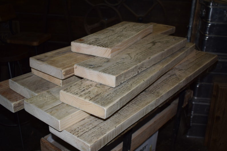 May include: A stack of reclaimed wood planks, showing the natural grain and texture of the wood. The planks are weathered and have a light gray color.