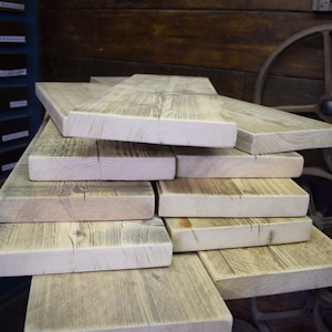 Op de afbeelding: Een stapel gerecyclede houten planken, die de natuurlijke nerf en knoesten van het hout laten zien. De planken zijn verweerd en hebben een lichtgrijze afwerking.