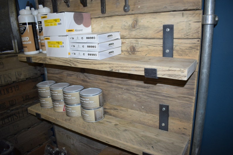 May include: Three wooden shelves with metal brackets, mounted on a rustic wood wall. The shelves are filled with various products, including bottles of liquid adhesive, boxes of sandpaper, and cans of wood wax.