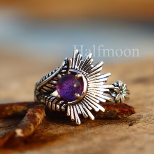 May include: A silver ring featuring a central purple gemstone, encircled by a sunburst pattern. The ring showcases detailed craftsmanship and a small floral element. It rests on a wooden surface.
