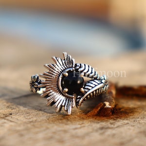 May include: A silver ring with a black gemstone centre, surrounded by a sunburst design. The ring features leaf and floral accents on the band. The ring is displayed on a wooden surface.