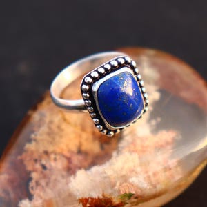Puede incluir: Un anillo de plata con una piedra preciosa cuadrada de color azul oscuro. La piedra preciosa está engastada en un bisel de plata con un borde de cuentas. El anillo se muestra sobre una superficie de piedra marrón y naranja.