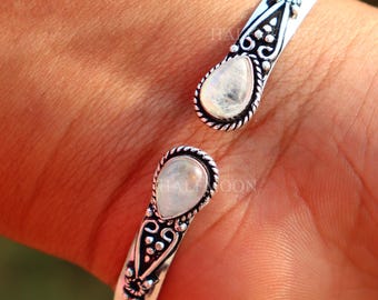 May include: A silver cuff bracelet featuring two teardrop-shaped moonstone cabochons at the open ends. The metal band displays intricate oxidized scrollwork and beaded detailing surrounding each stone, creating a textured, metallic finish.