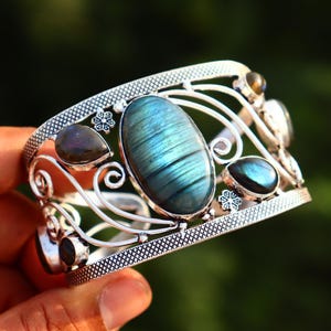 May include: A silver cuff bracelet featuring a large, oval-shaped, iridescent blue labradorite gemstone at the center, flanked by smaller teardrop-shaped labradorite stones and intricate metal scrollwork with small flower accents.