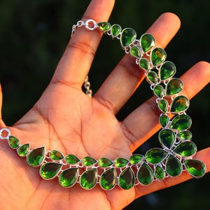 May include: A silver necklace featuring a series of vibrant green, teardrop-shaped gemstones. The gemstones are set in a silver metal frame and connected by a delicate chain. The necklace is displayed on an open hand against a blurred background.