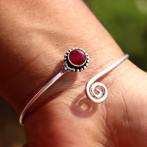 May include: A silver cuff bracelet with a large, round, deep red gemstone. The gemstone is set in a decorative bezel with a floral design. The bracelet has a spiral design on one end.