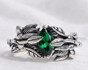 May include: A silver ring featuring a marquise-cut emerald green gemstone at the center. The band is designed with detailed metal leaves and small buds wrapping around the stone, creating a textured, organic appearance with a polished finish.