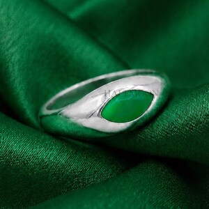 May include: A polished silver ring featuring a smooth, marquise-shaped green stone inlay set into the center of the band. The metal has a reflective, high-shine finish, and the green stone is vibrant and opaque, creating a contrast with the silver.