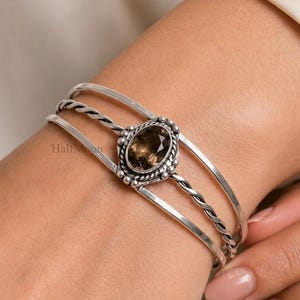 Puede incluir: Un brazalete de plata con tres bandas distintas, incluyendo un diseño central de cuerda trenzada. El centro está adornado con una piedra preciosa marrón de corte ovalado y facetado, engastada en un marco de plata con cuentas. La palabra HalfMoon es visible.