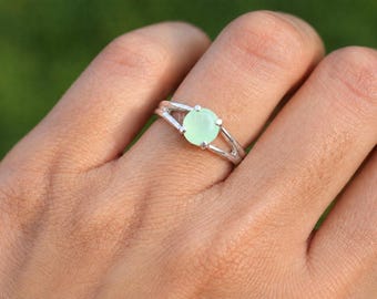 May include: A silver ring featuring a round, pale mint green cabochon stone set in a four-prong mount. The band is constructed from two thin, polished silver wires that split and rejoin at the base of the setting, creating an open design.