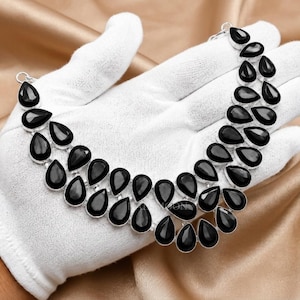 May include: A double-strand necklace featuring numerous black, pear-shaped stones set in silver-toned metal. The stones are arranged in two cascading rows, creating a curved, collar-like shape that follows the contour of the hand holding the piece.