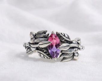 May include: A silver ring featuring a marquise-cut gemstone that transitions from pink to purple. The band is designed with detailed metal leaves and small berries wrapping around the center stone, creating a nature-inspired botanical aesthetic.
