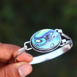 Peut inclure: Bracelet en argent avec une pièce centrale en forme d'ovale en coquille d'ormeau. La coquille présente des reflets bleus, verts et violets irisés, sertie dans une lunette en argent avec un motif de corde torsadée.