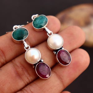 May include: Silver dangle earrings with three gemstones each. The top stone is a round, emerald green gem, followed by a white pearl, and a deep red oval gem at the bottom. The earrings are held in a hand.