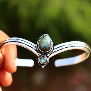 May include: A silver-toned cuff bracelet featuring a V-shaped design with a braided texture along the band. The centre is adorned with two teardrop-shaped labradorite stones, one larger than the other, set in decorative silver metal bezels.