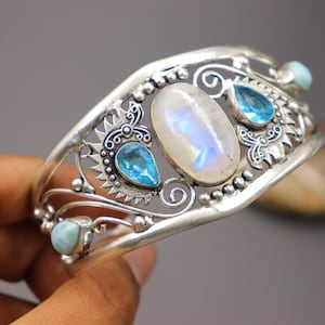 Pode incluir: Uma pulseira de punho prateada com uma grande pedra da lua oval e várias pedras preciosas azuis. A pulseira tem um design vazado com redemoinhos decorativos e detalhes de contas. As pedras preciosas são engastadas em engastes ornamentados.