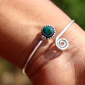 Könnte beinhalten: Silberfarbenes Armreif-Armband mit einem runden, dunkelgrünen Edelstein, der in einer Perlenfassung gefasst ist. Das Armband hat ein Spiraldesign an einem Ende und ein Blumenakzent in der Nähe des Edelsteins.