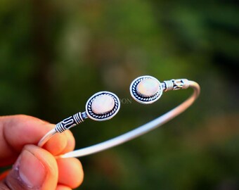 May include: An open-ended silver cuff bracelet featuring two oval-shaped, iridescent white stones set in decorative, twisted metal frames. The band displays intricate, etched detailing near the stone settings, providing a textured appearance.