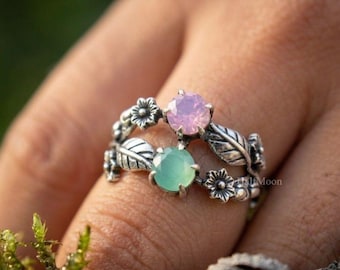 May include: A silver-toned ring featuring two stacked bands with floral and leaf motifs. The top band holds a round, faceted light pink stone, while the bottom band features a round, faceted mint green stone. The metal has an oxidized finish.