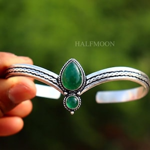 Può includere: Un bracciale a fascia in argento con un design a forma di V e una trama a corda intrecciata lungo la banda. Il centro è decorato con una pietra verde a goccia sopra una pietra verde rotonda. Il testo HALFMOON è visibile sullo sfondo.