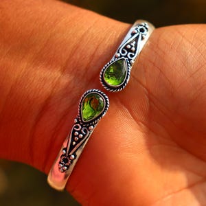 Puede incluir: Un brazalete de plata con dos piedras preciosas verdes en forma de pera. El brazalete presenta intrincados detalles de filigrana y está diseñado para usarse en la muñeca. Las piedras verdes son el punto focal de la joya.