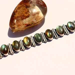 Puede incluir: Un brazalete de plata con piedras preciosas de labradorita de forma ovalada. El brazalete tiene un acabado de plata pulida y está acentuado con pequeñas cuentas de plata.