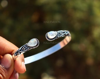 May include: A silver cuff bracelet featuring two teardrop-shaped moonstones at the open ends. The metal band displays intricate oxidized detailing and decorative patterns. The word HALFMOON is printed in small black letters on the bracelet surface.