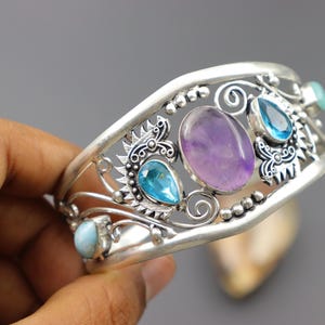 May include: Silver cuff bracelet featuring an oval amethyst, teardrop-shaped blue topaz, and other gemstones. The bracelet has an openwork design with decorative elements and is approximately 2 inches wide.