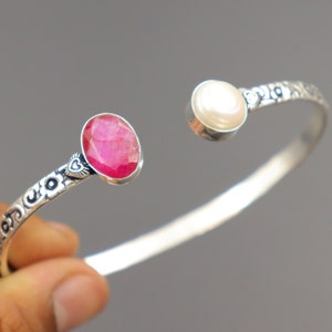 Könnte beinhalten: Ein silbernes Armreif-Armband mit einem rosafarbenen Edelstein und einer weißen Perle. Das Armband hat ein florales Design.