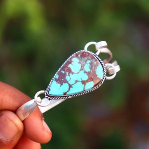 May include: A silver cuff bracelet featuring a central teardrop-shaped turquoise stone with brown matrix veining. The stone is set in a twisted rope border, and the bracelet band connects to the setting with circular silver loops on each side.