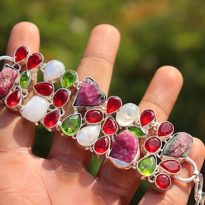 May include: A silver bracelet featuring a variety of colorful gemstones. The bracelet includes red, green, white, and pink stones in various shapes, including teardrops and irregular cuts. The bracelet is held in a hand against a blurred green background.