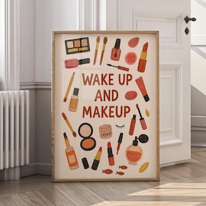Op de afbeelding: Een ingelijste print met illustraties van make-up items in oranje, rode en bruine tinten. De woorden "WAKE UP AND MAKEUP" staan in het midden. Het kunstwerk bevat afbeeldingen van lippenstift, oogschaduw en parfum.