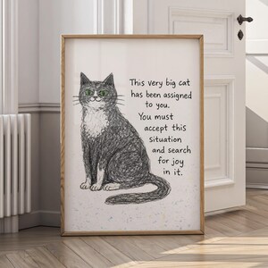 May include: A framed art print featuring a sketched black and white cat with green eyes. Text on the print reads: "This very big cat has been assigned to you. You must accept this situation and search for joy in it."