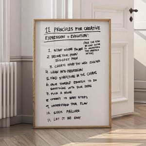May include: A framed whiteboard with handwritten text detailing "11 Principles for Creative Expression + Evolution." The text includes phrases like "Start Where You Are," "Become Your Own Biggest Fan," and "Seek Failure." The frame is a light brown color.